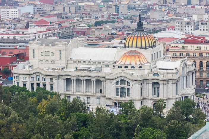 The Palacio de Bellas Artes seen slightly from above: a white marble palace with a colonnaded front and two domes, the larger one tiled in orange and yellow, with green trees along its front and the dense low roofline of the historic centre stretching away behind it.