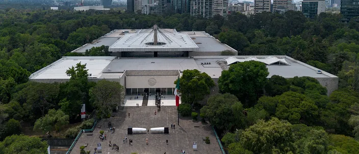 Aerial view of the National Anthropology Museum: a long low gray modernist block folded around an open courtyard, a wide paved forecourt with a red banner at the entrance and a few dozen visitors on it, dense green woodland on three sides and office towers on the skyline behind.