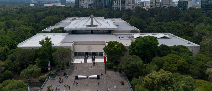 Aerial view of the National Anthropology Museum: a long low grey modernist block folded around an open courtyard, a wide paved forecourt with a red banner at the entrance and a few dozen visitors on it, dense green woodland on three sides and office towers on the skyline behind.