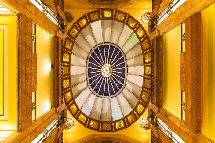 Looking straight up into the dome of the Palacio de Bellas Artes: an oval stained-glass lantern in blue and cream at the centre, ringed by amber glass panels and gilded coffering, with four marble piers rising into the corners of the frame.