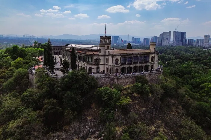 Chapultepec Castle from the air: a long cream palace with a square corner tower and a glazed upper terrace standing on a wooded rock outcrop, small figures along its terrace wall, dense woodland falling away below it and the city's towers and distant mountains on the horizon under a blue sky with white cloud.
