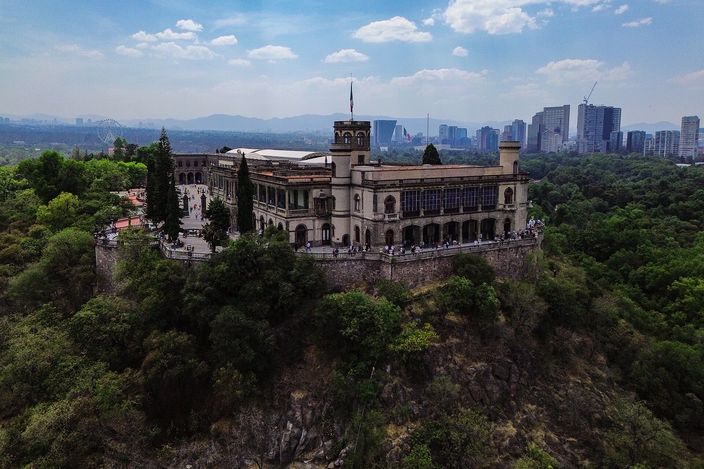 Chapultepec Castle from the air: a long cream palace with a square corner tower and a glazed upper terrace standing on a wooded rock outcrop, small figures along its terrace wall, dense woodland falling away below it and the city's towers and distant mountains on the horizon under a blue sky with white cloud.