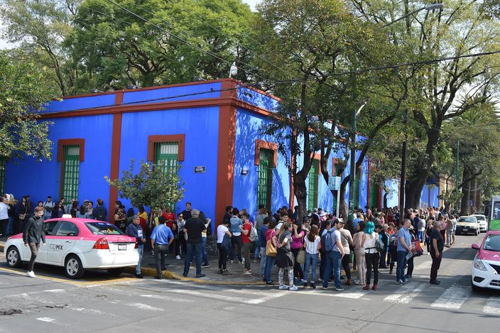 The corner of the Casa Azul in Coyoacan: cobalt blue walls with rust-red trim and green shuttered windows, a line of visitors waiting along the pavement under tall trees, and a pink-and-white Mexico City taxi at the kerb in the foreground.
