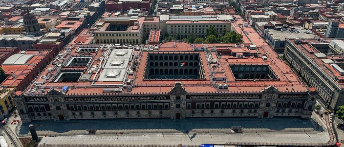 Aerial view of the Palacio Nacional filling one side of the Zocalo: a long two-storey facade of dark red stone with three portals under a red-tiled roof, its inner courtyards open to the sky, the grey flagstones of the square along the bottom edge and the dense low roofs of the historic centre stretching away behind.