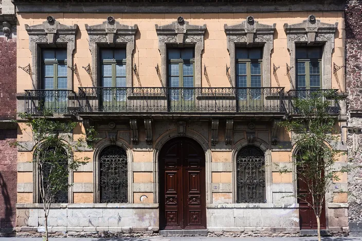 A two-story ochre-and-cream townhouse facade on cobbled Calle Mesones in the historic center: tall shuttered windows with a first-floor iron balcony, carved stone lintels and a dark wooden double door flanked by arched windows.