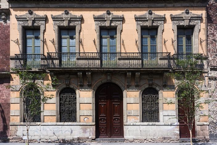 A two-storey ochre-and-cream townhouse facade on cobbled Calle Mesones in the historic centre: tall shuttered windows with a first-floor iron balcony, carved stone lintels and a dark wooden double door flanked by arched windows.