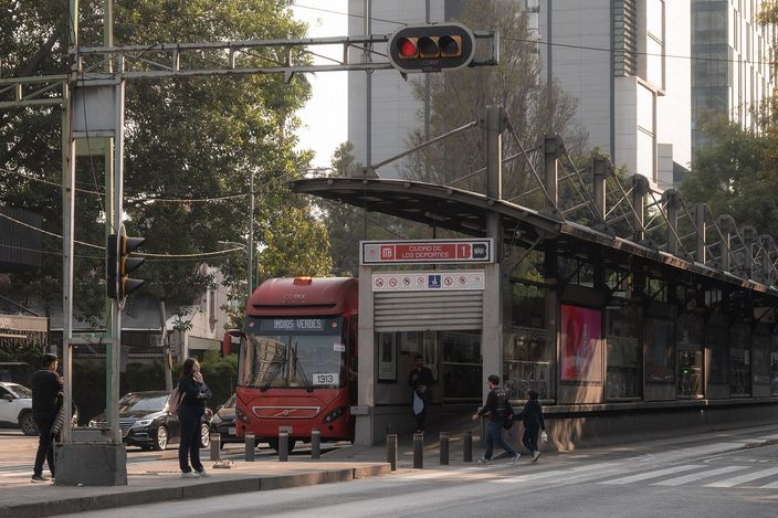 A red articulated Metrobús pulls into a median BRT station platform on a wide Mexico City avenue, with commuters crossing, traffic signals overhead and glass office towers behind trees