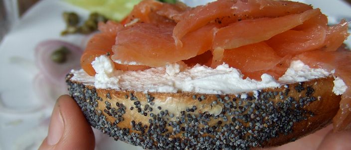 A halved poppy-seed Montréal bagel held in a hand, spread with cream cheese and topped with thin slices of smoked salmon, showing the dense ring and crisp seeded crust.
