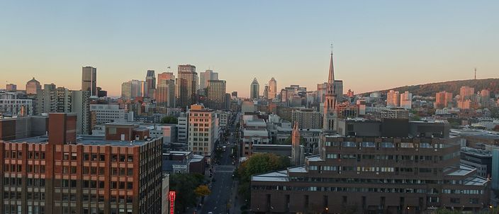 Montréal downtown skyline at sunrise seen from above, clusters of towers with Mount Royal's wooded slope and the illuminated cross rising to the right, a church spire mid-frame and Rue Saint-Denis running toward the core