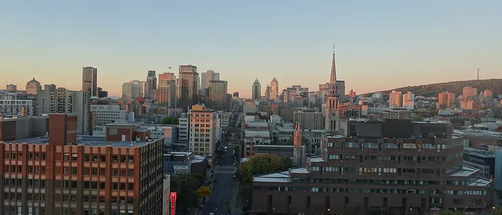 Montréal downtown skyline at sunrise seen from above, clusters of towers with Mount Royal's wooded slope and the illuminated cross rising to the right, a church spire mid-frame and Rue Saint-Denis running toward the core
