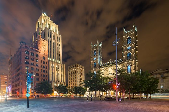 Place d'Armes at night, the twin grey stone towers of Notre-Dame Basilica floodlit on the right, the stepped art deco Aldred Building rising on the left, and the empty paved square in front of them