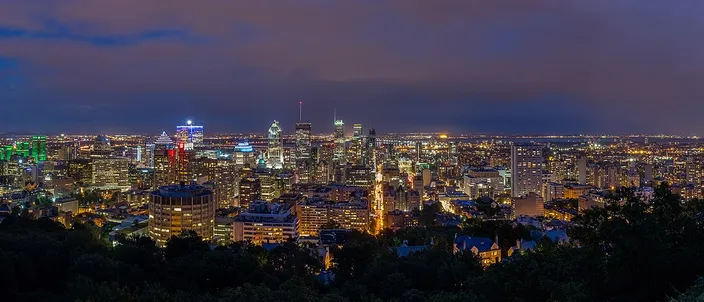 Night panorama of Montréal seen from the wooded slope of Mount Royal, a lit cluster of downtown towers across the center, a floodlit church spire on the ridge below them, and the low grid of the city spreading out to a dark horizon
