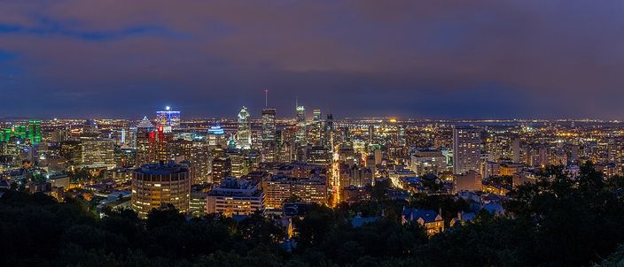 Night panorama of Montréal seen from the wooded slope of Mount Royal, a lit cluster of downtown towers across the centre, a floodlit church spire on the ridge below them, and the low grid of the city spreading out to a dark horizon