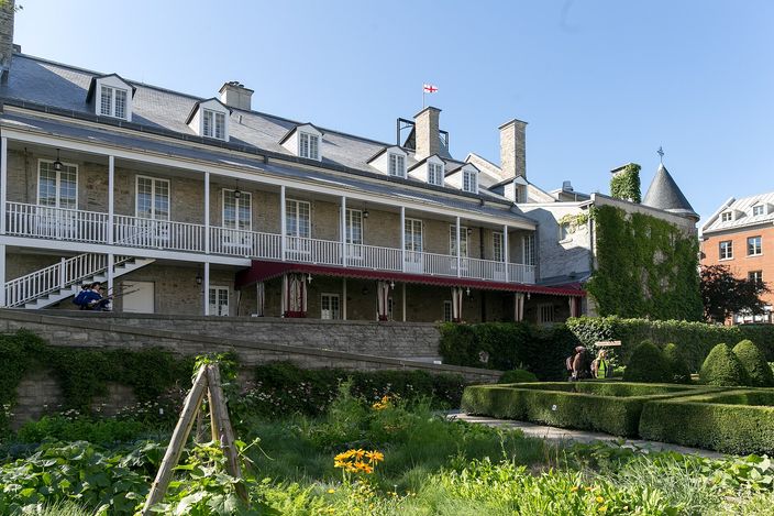 A long two-storey stone house with dormer windows, a wooden verandah and red awnings, seen across a terraced formal garden of clipped hedges and vegetable beds, with a small round turret to the right under a clear blue sky.
