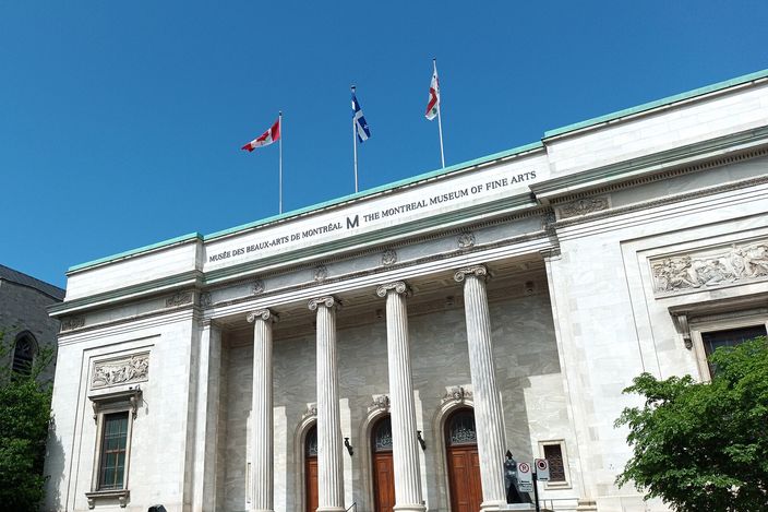 The white marble portico of a museum with four tall Ionic columns above a wide staircase, three flags flying from the roofline and the museum's bilingual name lettered along the frieze, against a clear blue sky.