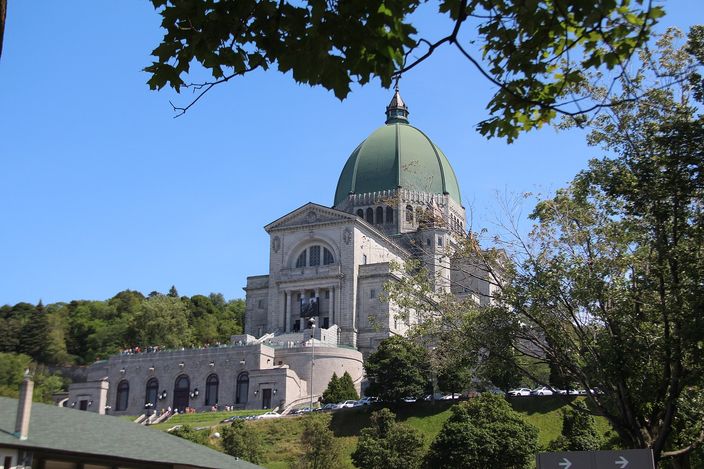 A large grey stone basilica with a green copper dome and a columned front standing on a wooded hillside above wide terraces and a long staircase, framed by tree branches, under a clear blue sky.