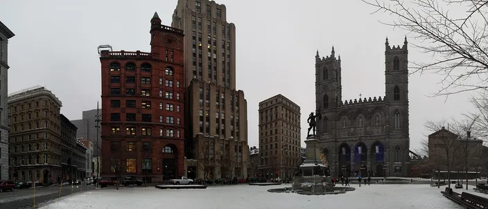 A wide paved square under snow, the twin gray stone towers of a Gothic church on the right, a bronze statue on a tall plinth in the middle, and a red sandstone Victorian block beside a stepped brown art deco tower on the left, under a flat gray sky.