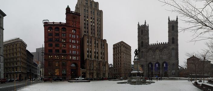 A wide paved square under snow, the twin grey stone towers of a Gothic church on the right, a bronze statue on a tall plinth in the middle, and a red sandstone Victorian block beside a stepped brown art deco tower on the left, under a flat grey sky.