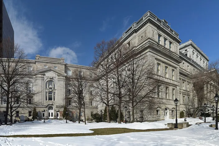 The gray-stone Second Empire Lucien-Saulnier building (old courthouse) in Vieux-Montréal under a clear blue winter sky, with bare trees and patches of snow on the lawn in front.