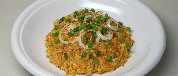 A mound of orange, paprika-tinged Obatzda cheese spread topped with raw onion rings and chopped spring onion, served on a white plate