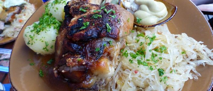 A whole roasted pork knuckle with dark, crisp, lacquered skin, served with a round white dumpling, shredded sauerkraut and dark gravy on a plate