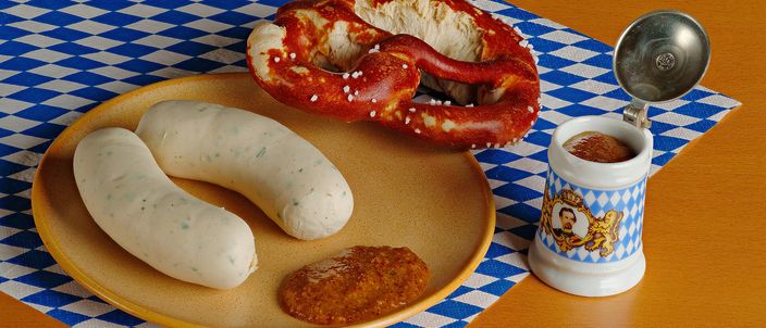 Two pale Weißwurst sausages on a plate with a soft pretzel and a dab of sweet brown mustard, laid on a blue-and-white Bavarian check cloth