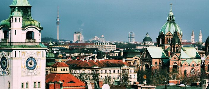 Munich rooftops in warm light: green copper church towers and red-tiled roofs in the foreground, the slim Olympiaturm and distant twin-domed churches on the skyline behind.
