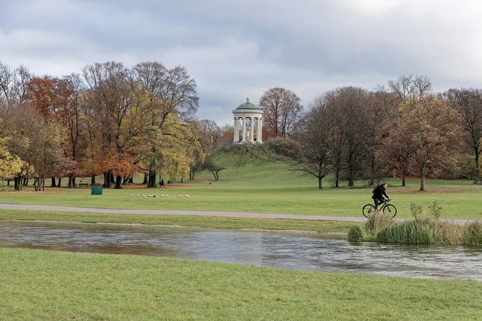 A round white colonnaded temple with a green dome standing on a grassy mound, ringed by beeches in orange and yellow autumn leaf, with a wide mown meadow, a stream in the foreground and a cyclist riding the path across it.