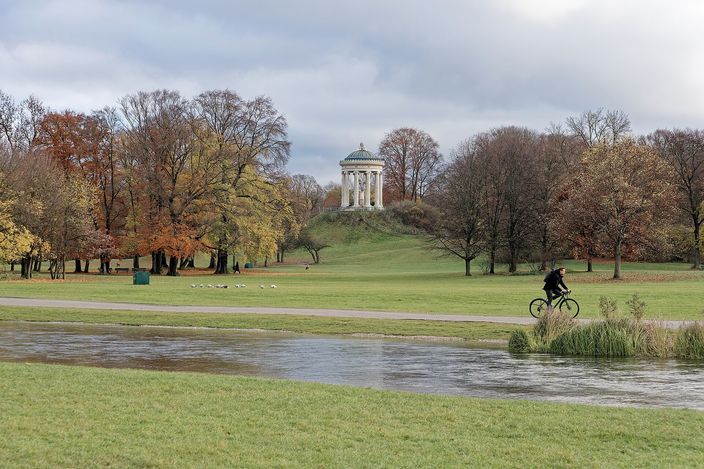 A round white colonnaded temple with a green dome standing on a grassy mound, ringed by beeches in orange and yellow autumn leaf, with a wide mown meadow, a stream in the foreground and a cyclist riding the path across it.