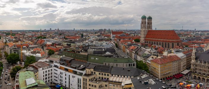 Munich's old town from a church tower: red-tiled roofs running to the horizon under heavy grey cloud, the Frauenkirche's brick nave and twin green onion domes standing above them on the right, and the New Town Hall's pale gothic arcade with red parasols and market stalls at the far right edge.