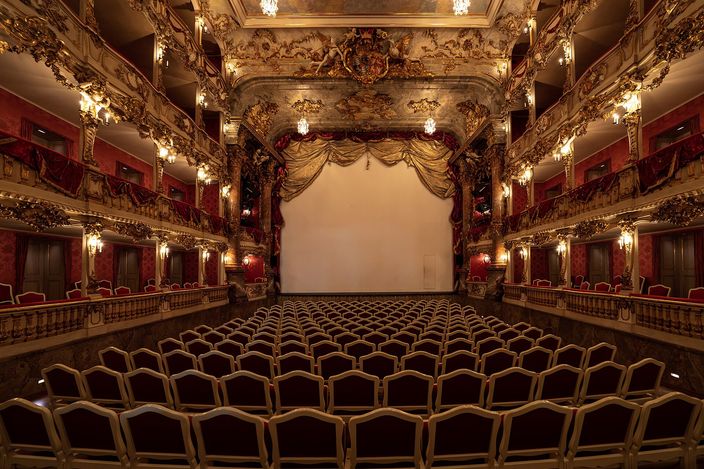 The Cuvilliés Theatre seen from the back of the stalls: four tiers of carved and gilded rococo boxes curving around a small auditorium, red damask panels between the piers, candle-shaped lamps on every column, and rows of empty pale chairs running forward to a plain lowered curtain.