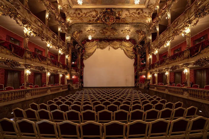 The Cuvilliés Theatre seen from the back of the stalls: four tiers of carved and gilded rococo boxes curving around a small auditorium, red damask panels between the piers, candle-shaped lamps on every column, and rows of empty pale chairs running forward to a plain lowered curtain.