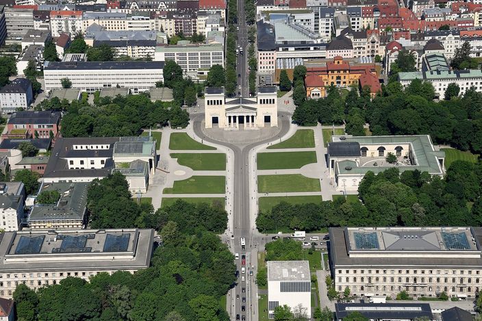 Königsplatz photographed from the air in summer: a Doric gateway closing the far end of a wide rectangle of lawns and pale paved paths, a low colonnaded building on either side, a tree-lined street and traffic crossing the near edge, and the roofs and courtyards of Maxvorstadt spreading away on all sides.