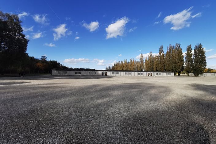 The empty gravel roll-call square at the Dachau memorial under a blue sky with scattered white cloud: a long low white building with regular barred openings runs across the far side, a line of poplars in autumn colour stands to the right, and the whole foreground is bare ground.