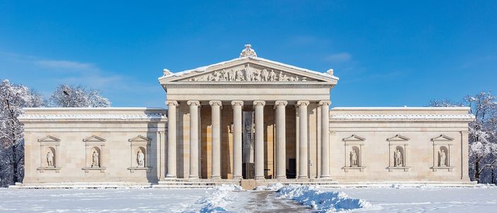 The Glyptothek on Königsplatz after snow: a low neoclassical museum with an eight-column Ionic portico and a sculpted pediment, statues standing in niches along the flanking wings, snow-laden trees behind it and untrodden snow across the ground in front, under a clear blue sky.