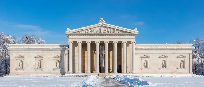The Glyptothek on Königsplatz after snow: a low neoclassical museum with an eight-column Ionic portico and a sculpted pediment, statues standing in niches along the flanking wings, snow-laden trees behind it and untrodden snow across the ground in front, under a clear blue sky.