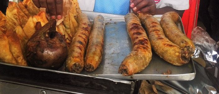 Five grilled mutura sausages on a metal tray at a street stall, beside fried snacks and a piece of grilled offal, with the vendor's hands above