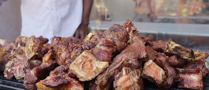A heap of meat pieces browning on a charcoal grill, with a cook in a white coat standing behind it