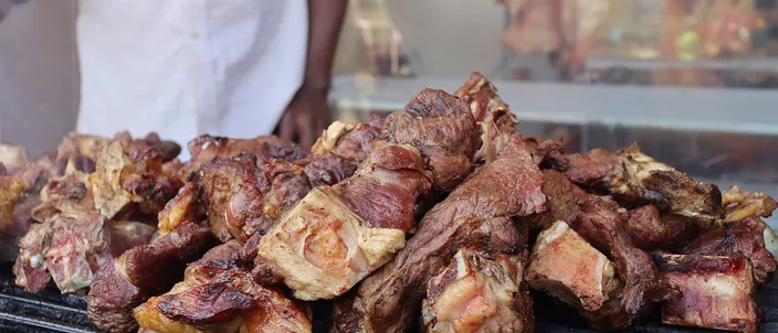A heap of meat pieces browning on a charcoal grill, with a cook in a white coat standing behind it