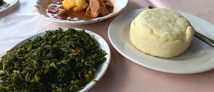 A pale dome of ugali on a plate beside a dish of cooked greens, on a restaurant table