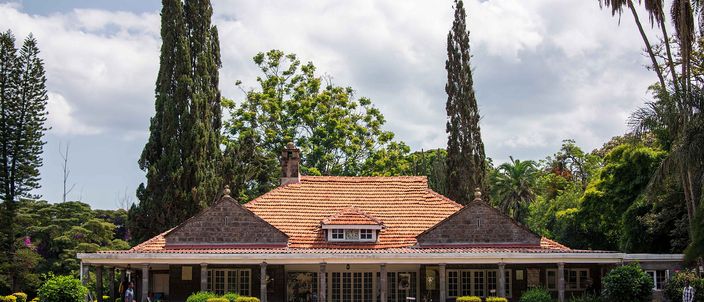 A single-storey stone bungalow with a steep red-tiled roof and a long colonnaded verandah, standing behind a wide mown lawn between tall dark cypresses and palms under a blue sky with white cloud.
