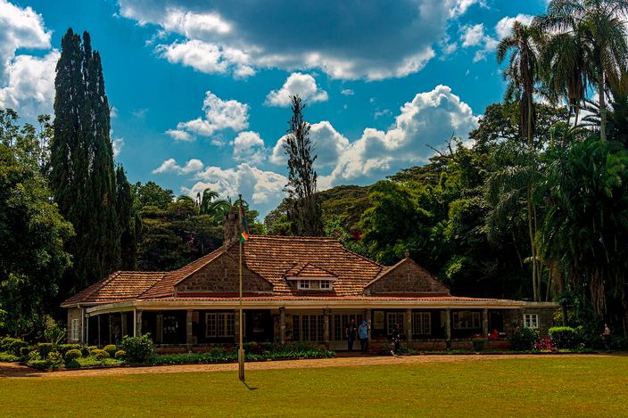 A single-storey colonial bungalow with a red-tiled roof and verandah, set among tall cypress and palm trees behind a broad green lawn under a bright cloud-filled sky
