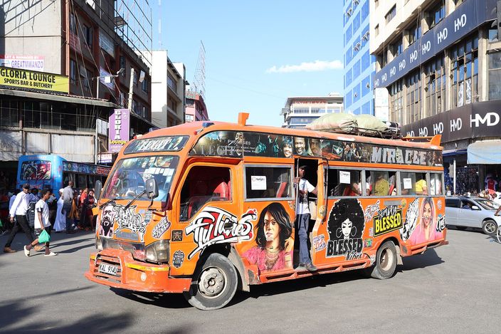 A brightly painted orange matatu minibus covered in graffiti-style portraits and slogans, packed with passengers, on a busy Nairobi city-centre street lined with shops and pedestrians