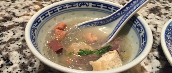 A blue-and-white porcelain bowl of duck blood and vermicelli soup on a granite counter, with clear glass vermicelli, cubes of dark duck blood, a patterned spoon resting in the bowl, and a small dish of chilli oil blurred in the background.