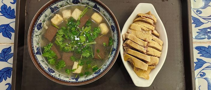 A bowl of duck blood and vermicelli soup beside a white dish of sliced pale ivory Nanjing salted duck, on a tray over blue-and-white tiled placemats.