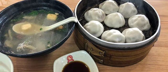 A steamer basket of plump, thick-skinned soup buns beside a bowl of duck blood and vermicelli soup and a small dish of dark vinegar, on a wooden restaurant table in Nanjing.