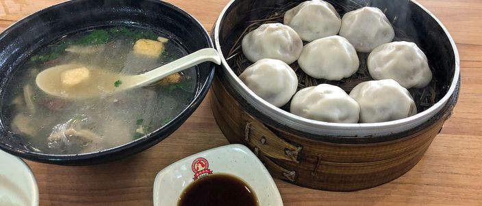A steamer basket of plump, thick-skinned soup buns beside a bowl of duck blood and vermicelli soup and a small dish of dark vinegar, on a wooden restaurant table in Nanjing.