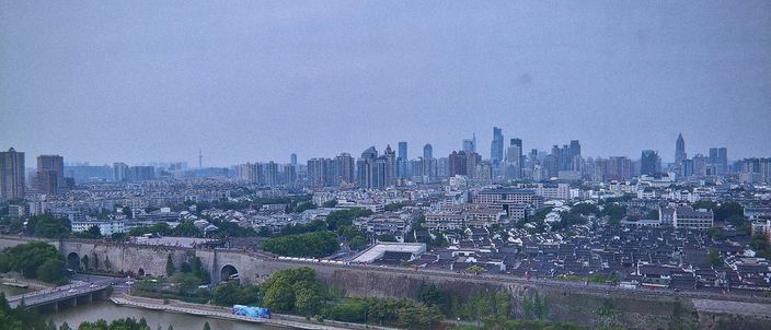 A hazy view over Nanjing from the Porcelain Pagoda: a stretch of the Ming city wall and a gate tower beside a canal in the foreground, ordinary old-town rooftops beyond, and the modern high-rise skyline further back.