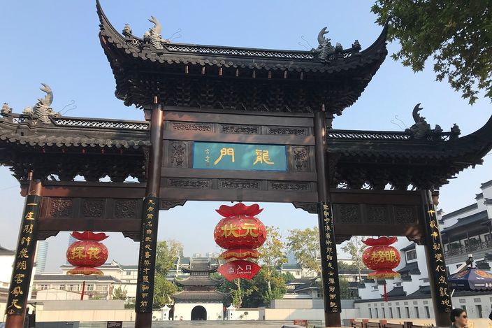 A dark-wood ceremonial paifang gateway with a curved, tiled, upturned roofline hung with red lanterns, framing a view through to the Jiangnan Examination Hall's courtyard buildings beyond.