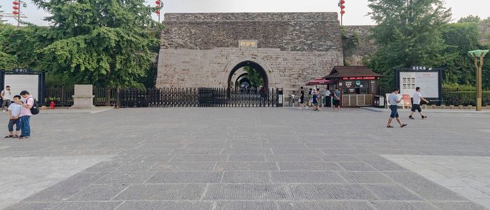 A wide stone plaza in front of Zhonghua Gate's Ming-dynasty wall, its single visible arch flanked by small red flags, with a scatter of visitors crossing the paving.
